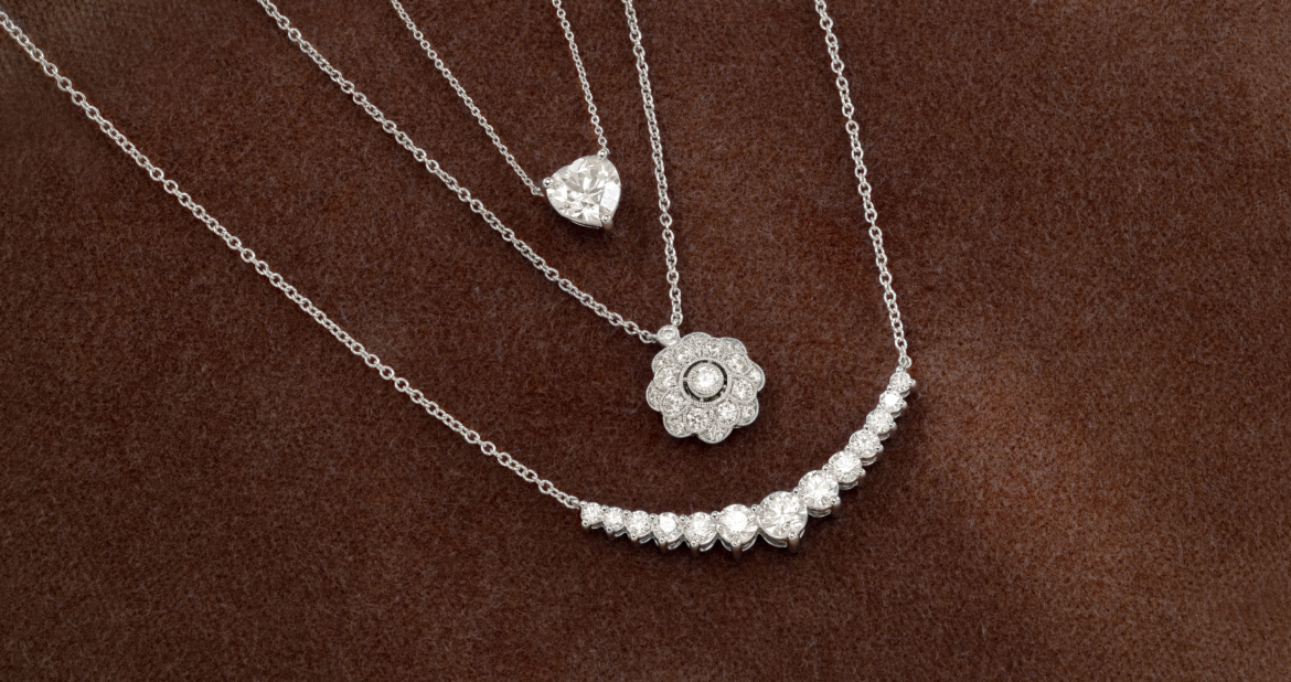 Three diamond necklaces on brown fabric - heart pendant, flower pendant, and graduated tennis necklace.