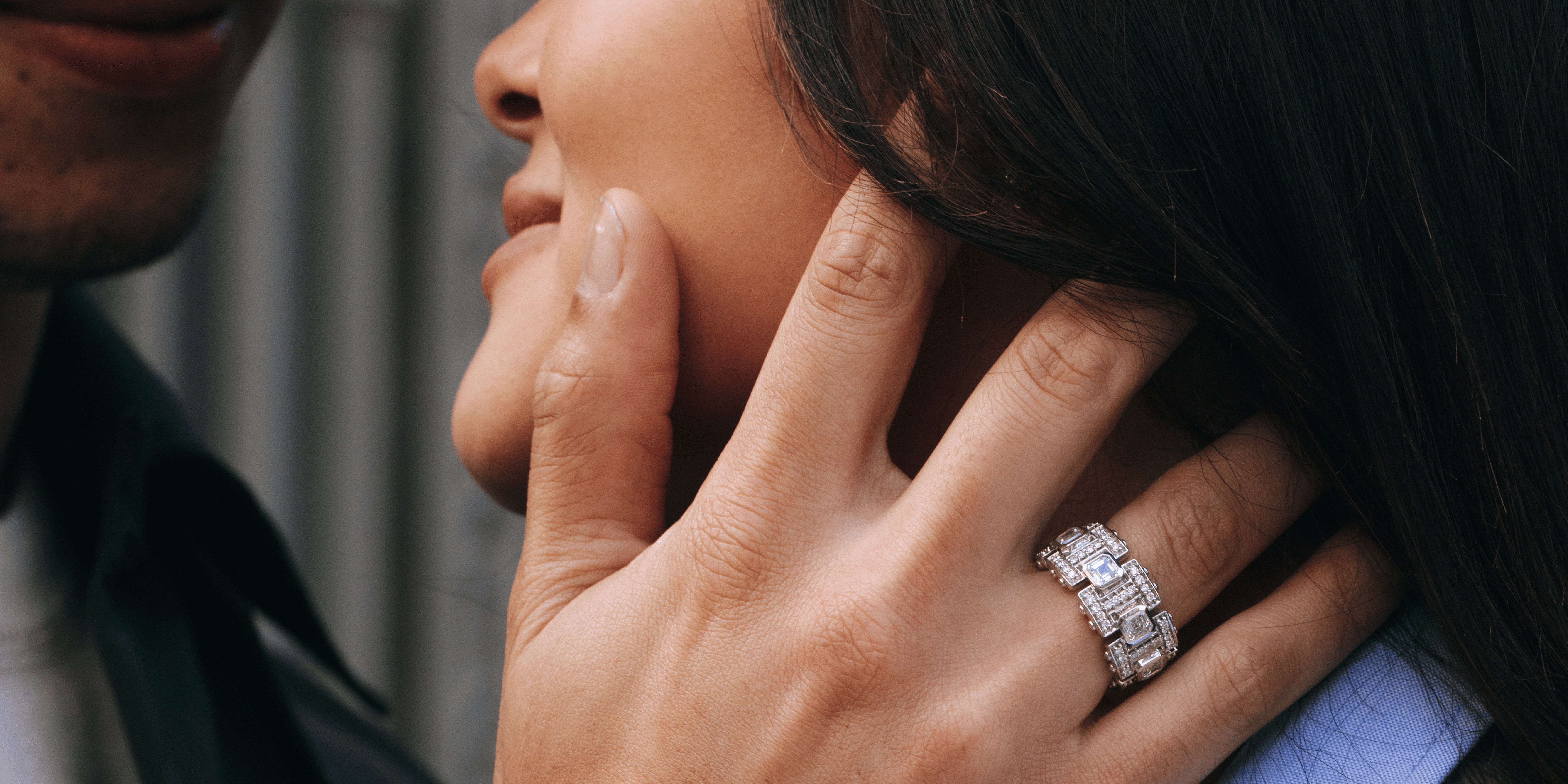 Close-up of a couple embracing, with the woman's hand gently resting on the man's face, showcasing a bold, multi-row diamond ring on her finger.