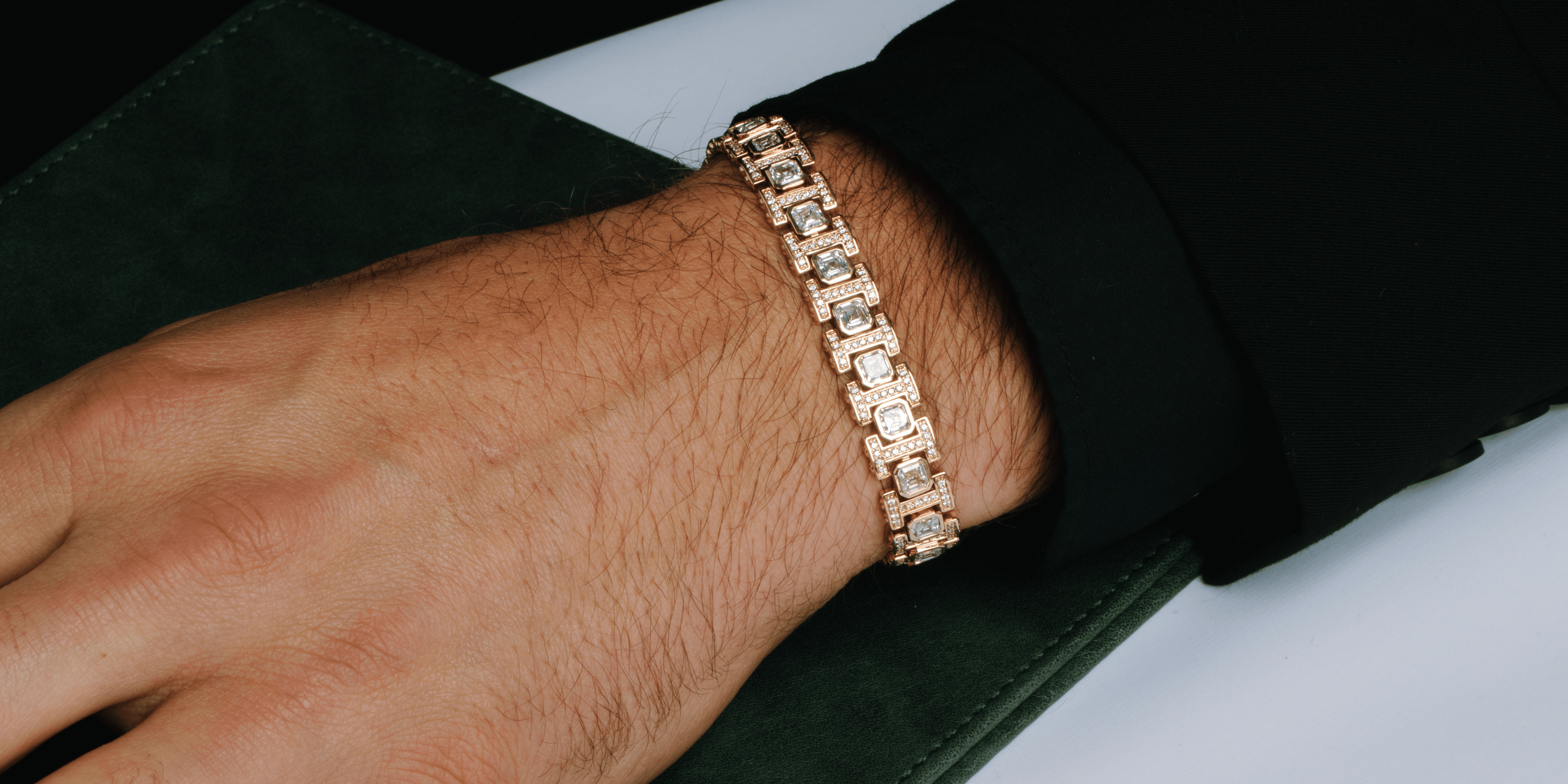 Close-up of a man's wrist wearing a gold bracelet with rectangular diamond accents, paired with a black sleeve and resting on a dark green textured surface.