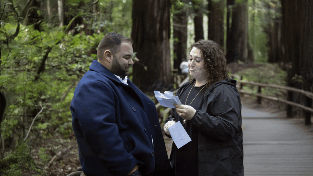 A man and woman stand together on a wooded path in Muir Woods. The woman is reading a note, visibly emotional, while holding several letters. The man looks on warmly, creating an intimate and heartfelt moment surrounded by tall redwoods.